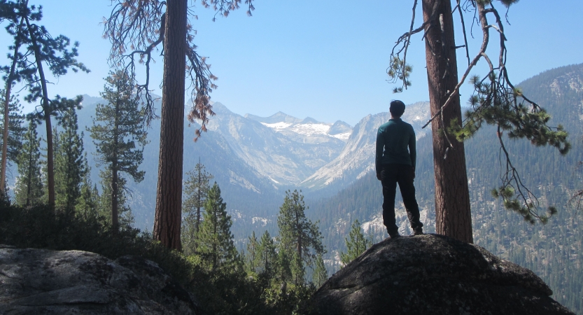 A person stands on a rock facing away from the camera, looking out over a vast mountainous landscape. There is snow on the mountains in the distance. 